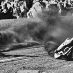 Two speedway motorcycles slide through a dirt turn, kicking up smoke