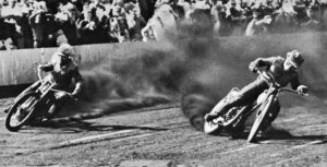 Two speedway motorcycles slide through a dirt turn, kicking up smoke